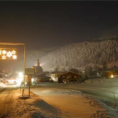 Eine verschneite Landschaft bei Nacht mit festlicher Beleuchtung. Im Hintergrund sind Häuser und eine Kirche zu sehen, umgeben von schneebedeckten Bäumen.