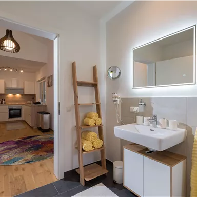 A modern bathroom with a stylish sink and a lit mirror. In the background, there is an open kitchen with carpet flooring.