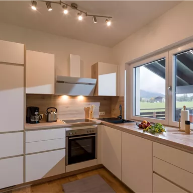 A modern kitchen with white furniture and a large window front. The room is bright and inviting, with a view of the landscape outside.