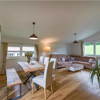 A modern and bright living room with a large dining table and a cozy couch. Large windows provide plenty of daylight and offer a view of the greenery.