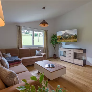 A cozy living room with a large window and a view of the green landscape. The decor consists of a beige couch, a wooden table, and a television on the wall.