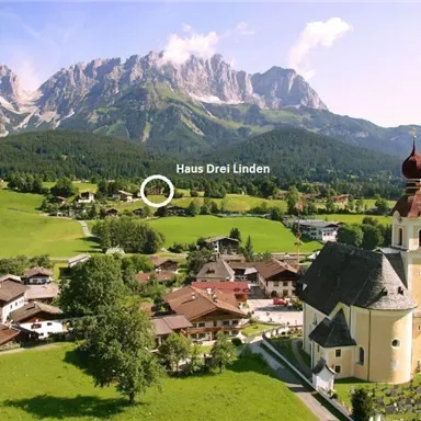 Eine malerische alpine Landschaft mit grünen Wiesen und majestätischen Bergen. Im Vordergrund steht eine schöne Kirche und traditionelle Häuser.