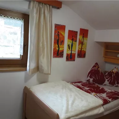 Ein gemütliches Schlafzimmer mit einem Einzelbett und bunten Wandbildern. Das Zimmer hat ein Fenster mit Blick nach draußen und eine freundliche, helle Atmosphäre.