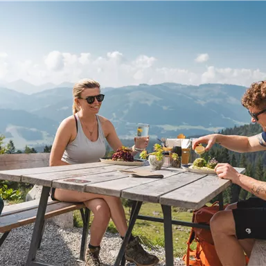 Ein Paar genießt eine Mahlzeit an einem Tisch in den Bergen. Im Hintergrund sind majestätische Berge und ein klarer blauer Himmel zu sehen.