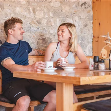 Ein Paar sitzt am Tisch in einem gemütlichen Raum mit Holzverkleidung. Sie lächeln einander an und genießen Getränke.
