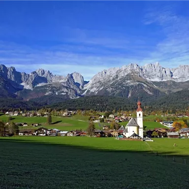 A picturesque landscape with high mountains and a small village. In the foreground stands a church, surrounded by green meadows and traditional houses.
