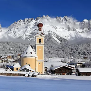 A picturesque winter landscape with snow-covered mountains. In the foreground stands a yellow church with a distinctive tower.