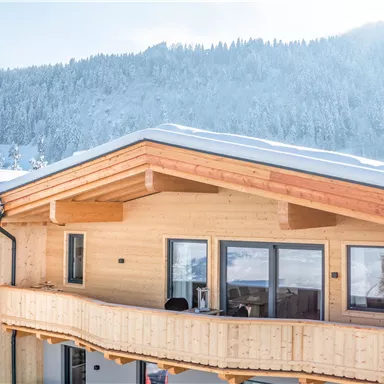 Ein modernes Gebäude aus Holz mit einer großen Terrasse. Im Hintergrund sind schneebedeckte Berge und ein klarer Himmel zu sehen.
