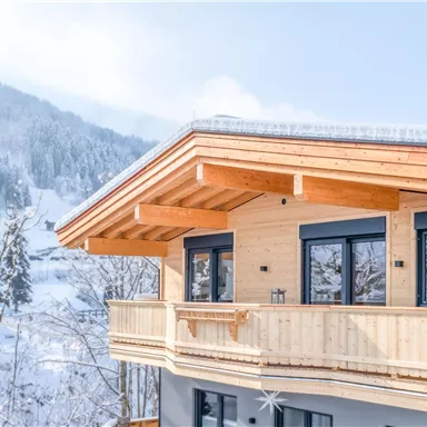 Ein modernes Holzhaus in den Bergen, umgeben von schneebedeckten Bäumen. Der Himmel ist klar und blau, was eine ruhige Winteratmosphäre schafft.