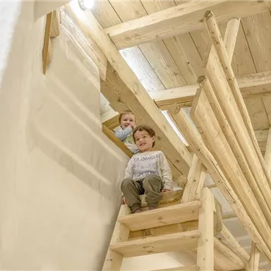 Zwei Kinder spielen in einem Holzhaus. Eines sitzt auf einer Treppe und das andere schaut aus einem Fenster.