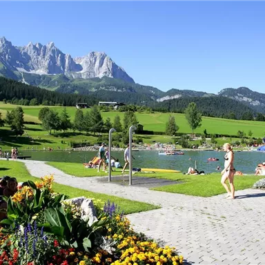 A beautiful lake surrounded by green meadows and mountains. People enjoy the sun and relax on loungers.