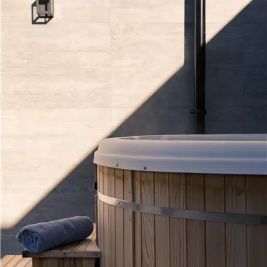A wooden hot tub stands against a wall of gray tiles. A towel lies on the wooden bench next to it.