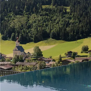 Ein malerisches Dorf mit einer Kirche, umgeben von grünen Wiesen und Wäldern. Im Vordergrund spiegelt sich das Wasser eines Pools.
