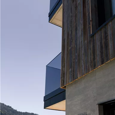 Ein modernes Gebäude mit Holz- und Glasdetails in einer bergigen Landschaft. Im Hintergrund sind sanfte Hügel unter klarem Himmel zu sehen.