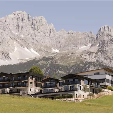 A modern-designed hotel building is set against an impressive mountain landscape. The surrounding nature is green and offers a peaceful atmosphere.