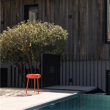 A modern outdoor area with a pool and an orange chair. In the background, there is a tree and a wooden facade of the house is visible.