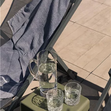 A deck chair with a blue towel and a small table. On the table are a carafe of water and two glasses.
