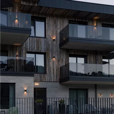 A modern building with a wooden facade and glass balconies. The facade is stylishly illuminated and has an elegant fence.