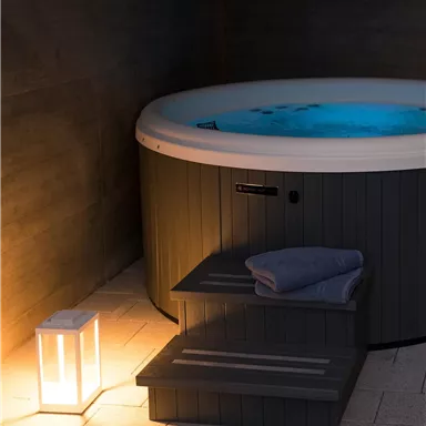 Ein Whirlpool in einem dimmbaren Raum, beleuchtet von einer kleinen Laterne. Neben dem Whirlpool liegen ein Handtuch und eine Treppe.
