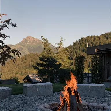 A campfire in a stone fire pit surrounded by nature. Mountains and trees can be seen in the background.