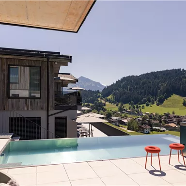 A modern terrace with an infinity pool and a view of the surrounding mountains. Red bar stools invite you to relax.