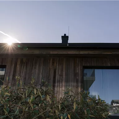 A modern wooden house with large windows. The sun shines over the roof, creating a warm atmosphere.