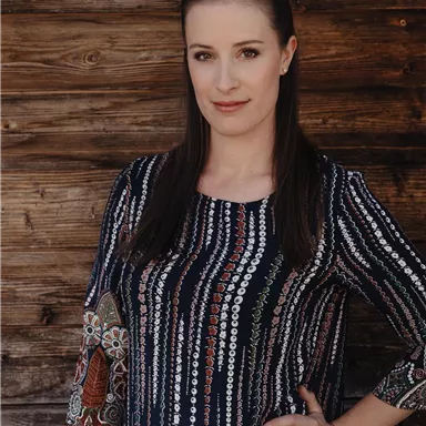 Eine Frau steht vor einer Holzwand und trägt ein gemustertes Oberteil. Sie hat lange Haare und schaut direkt in die Kamera.