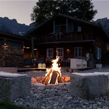 A cozy wooden house with a campfire in the foreground. In the background, mountains and a serene landscape can be seen.