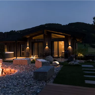 A cozy house outdoors at night, surrounded by a beautiful garden. A campfire flickers, creating a warm atmosphere.