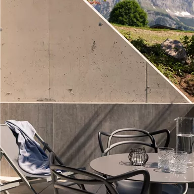 A modern terrace with a round table, chairs, and a lounge chair. In the background, mountains and a blue sky are visible.