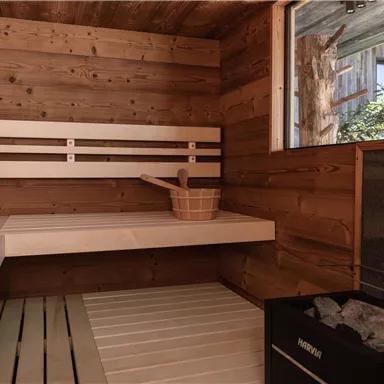 A cozy sauna with wood paneling and benches. On the left side, there is a bucket with a ladle and a stove for the stones.