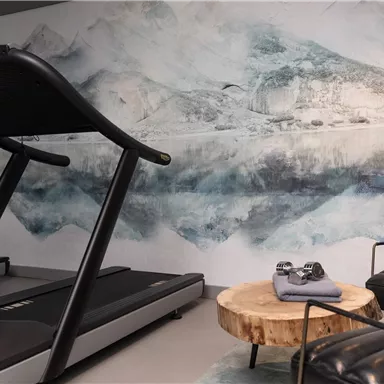 Ein moderner Fitnessraum mit zwei Laufbändern und einer stilvollen Holzbank. Im Hintergrund ist eine künstlerische Wandgestaltung in sanften Blau- und Grautönen.