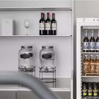 Eine moderne Bar mit Weinflaschen und Gläsern auf einem Regal. Neben einem Kühlschrank sind verschiedene Getränke in Flaschen sichtbar.