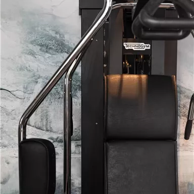 A modern fitness device with chrome-plated handles and a padded seat. The background features a minimalist wall design.