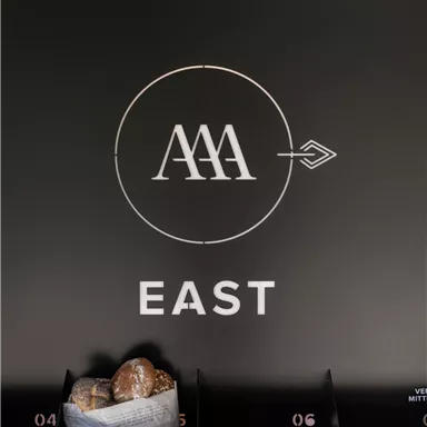 Ein schwarzes Schild mit dem Logo "AAA" und dem Wort "EAST". Darunter befinden sich Fächer mit einem Korb, der einige Brötchen enthält.