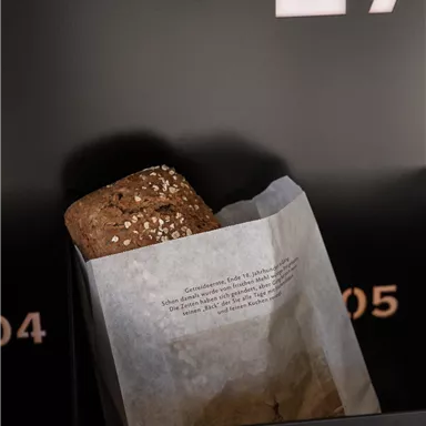 Ein leckeres Brötchen in einer Papierverpackung. Im Hintergrund sind schwarze Wände mit weißen Schriftzügen sichtbar.