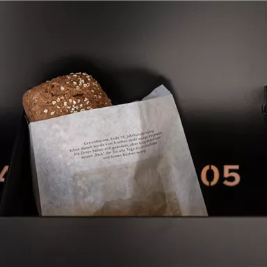 Ein Brötchen mit Sesam teilweise aus einer Papiertüte sichtbar. Im Hintergrund sind schwarze Wände mit Zahlen zu sehen.