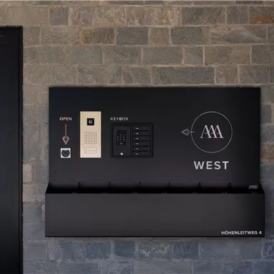 A modern mailbox with a black design on a stone wall. It features various compartments and functions for mail and keys.