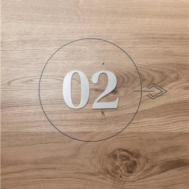 Eine helle Holzoberfläche mit der Zahl 02 und einem Pfeilsymbol. Die Zahl ist in der Mitte eines Kreises platziert.