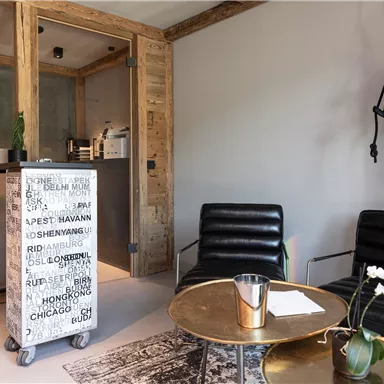 Ein modernes Wartezimmer mit Schwarzleder-Sesseln und einem runden Tisch. An der Wand hängt eine stilisierte Affenskulptur und der Raum hat eine gemütliche Atmosphäre.