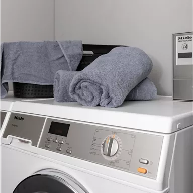 Eine moderne Waschmaschine mit grauen Handtüchern darauf. Im Hintergrund befindet sich ein schwarzer Behälter und eine Steuerungseinheit.