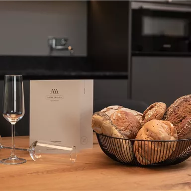 Ein Korb mit verschiedenen Brotsorten steht auf einem Holztisch. Daneben befinden sich zwei Gläser und ein Menü.