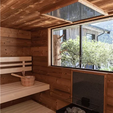 Eine gemütliche Sauna mit Holzverkleidung und einer Glastür. Im Hintergrund sind Bäume sichtbar, die durch das Fenster hineinblicken.