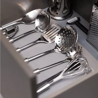 An open drawer with various stainless steel kitchen utensils. The utensils are neatly arranged and include spoons, a ladle, and a grater.