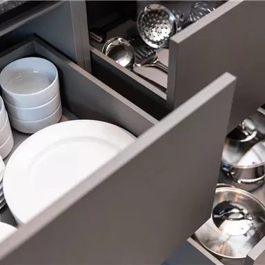 Two open drawers in a modern kitchen. The upper drawer contains white plates and bowls, while the lower drawer displays kitchen utensils and pots.