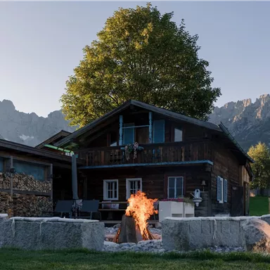 A cozy wooden house with a fire pit in the foreground. In the background, there are mountains and a clear sky.