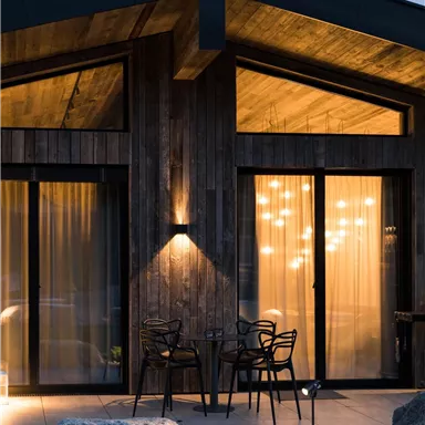 A modern wooden house with large windows and soft lighting. Outside, a few chairs and a table are placed on a terrace.