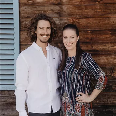 A man and a woman are standing next to each other in front of a wooden wall. Both are smiling and appear friendly.