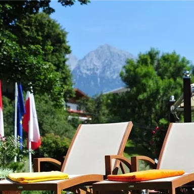 Two loungers with colorful cushions are in a garden. In the background, trees and mountains can be seen.