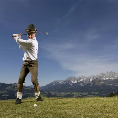 Ein Mann spielt Golf in traditioneller bayerischer Tracht. Im Hintergrund sind majestätische Berge und ein klarer Himmel zu sehen.